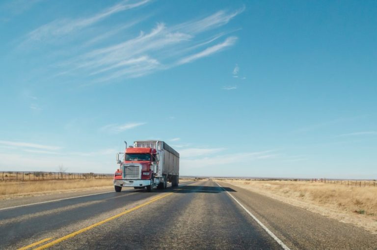 Red semi-truck hauling freight on an open two-lane highway through flat plains under a blue sky