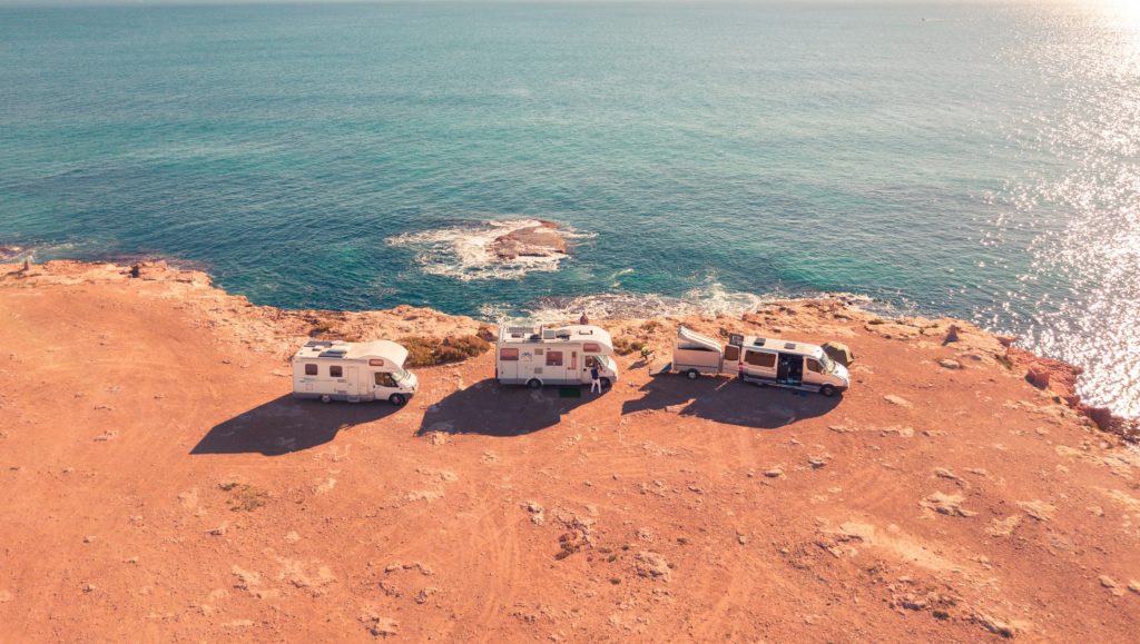 Aerial view of three RVs and a camper van parked on a coastal cliff overlooking turquoise water