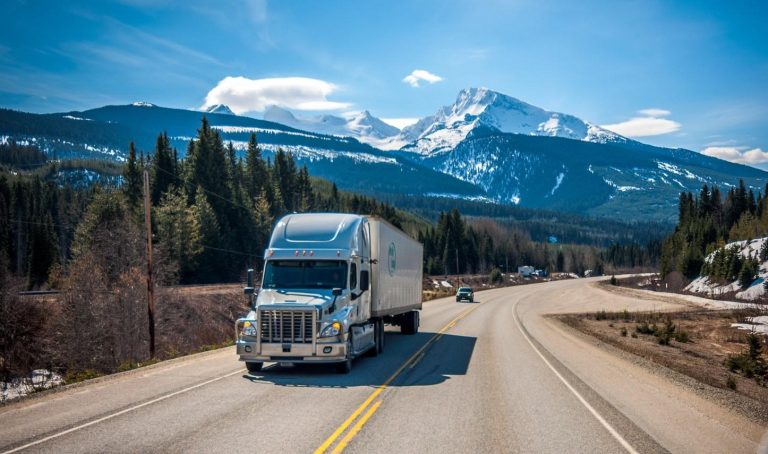 Semi-truck hauling a freight trailer on a mountain highway through forested terrain with snow-capped peaks, representing the modern trucking and transportation industry