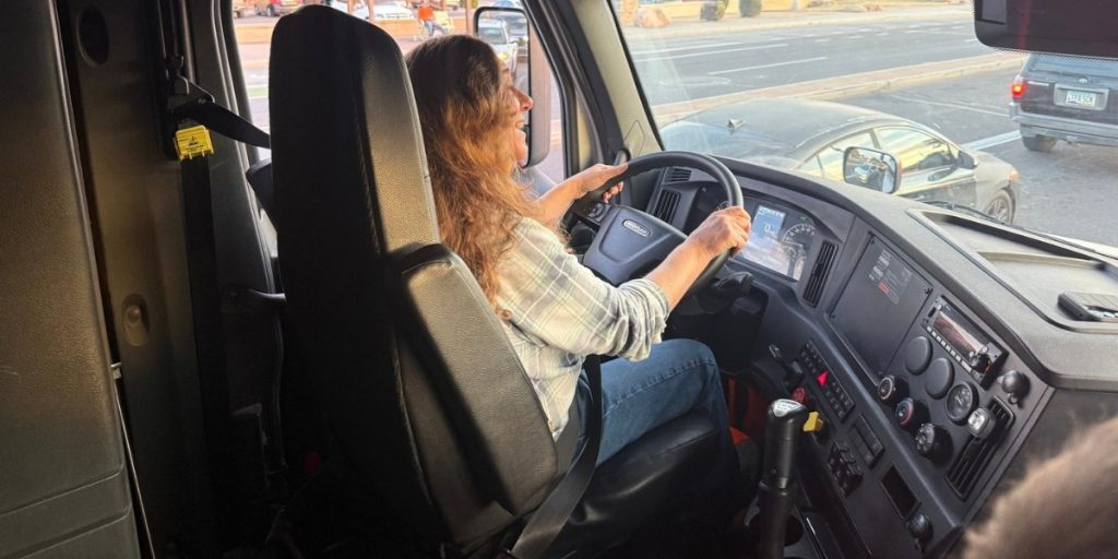 Woman truck driver behind the wheel of a semi-truck cab navigating city streets as an owner-operator