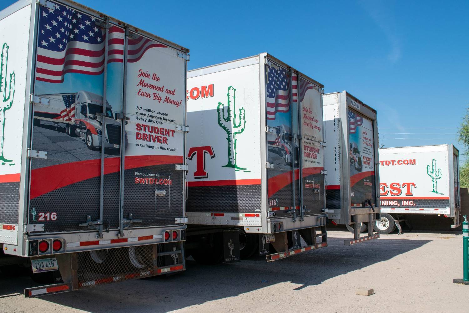Rear view of multiple Southwest Truck Driver Training semi-trailers parked in a lot, each wrapped with graphics featuring an American flag, and a cactus, under a clear blue sky.