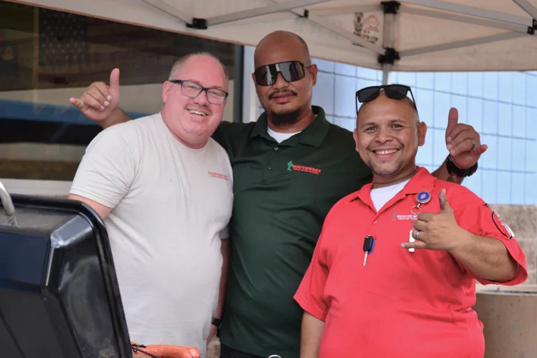 Three smiling Southwest Truck Driver Training staff members pose together beside a grill of hot dogs, two of them giving shaka hand signs.