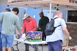 Attendees gather at a folding table set with condiments, utensils, and a large box of chips under a canopy tent