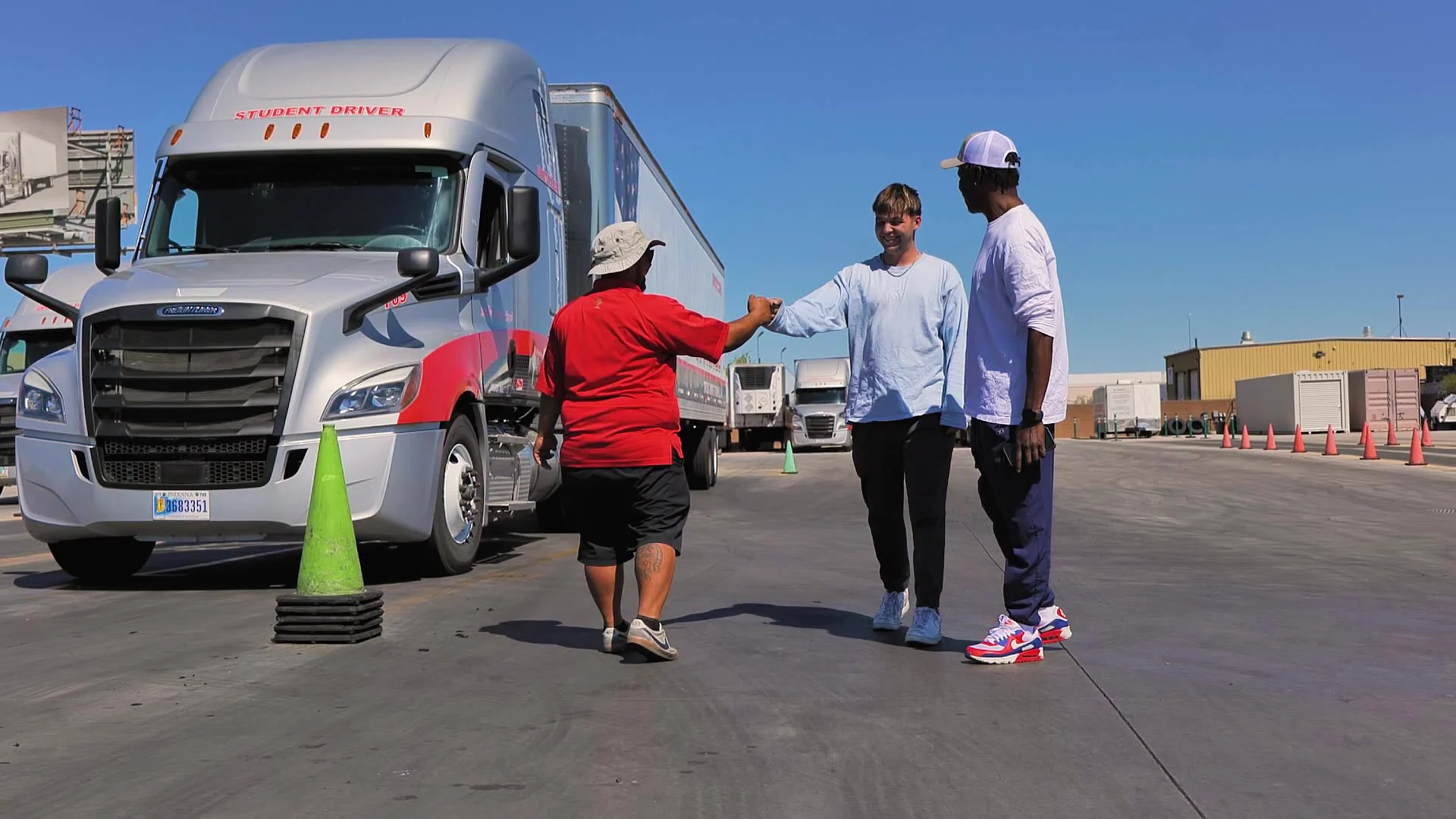 A smiling student in a light blue shirt fist-bumps an instructor in a red shirt and bucket hat on a truck driving range, with another student looking on beside a silver "Student Driver" semi truck and orange training cones.