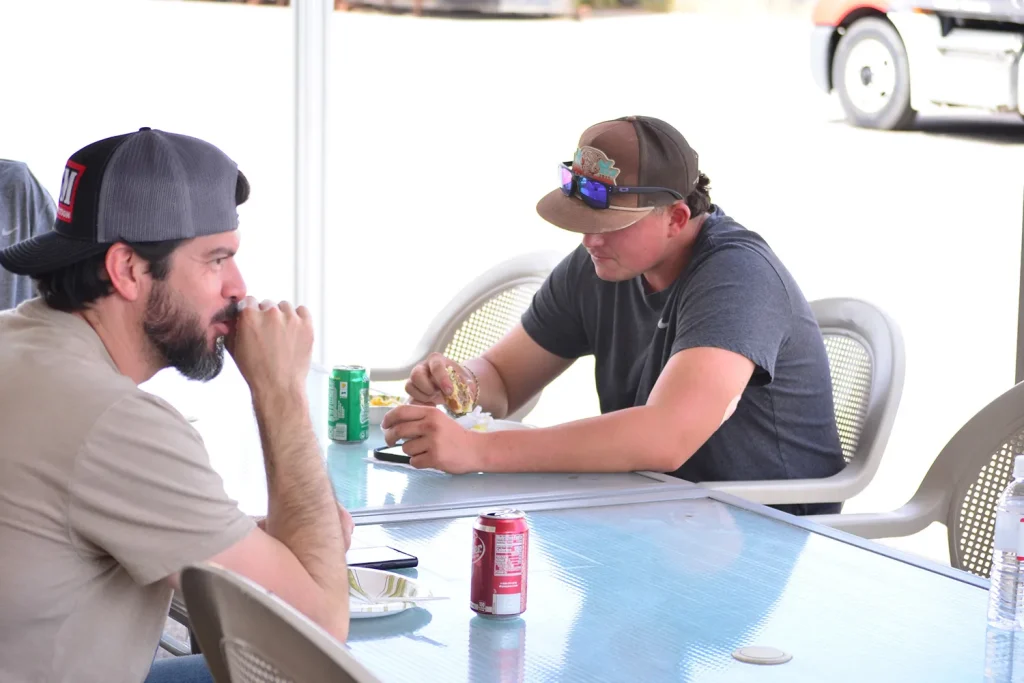 Two men in ball caps sit at a patio table with soda cans, one eating a burger and the other resting his chin on his hand in conversation.