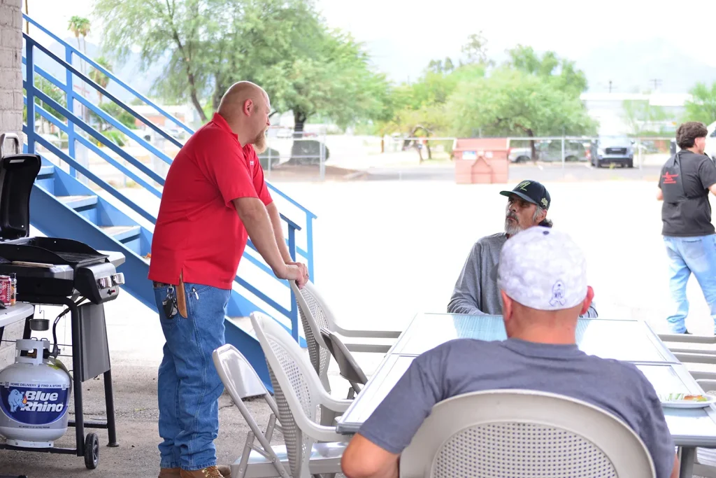A Southwest Truck Driver Training staff member in a red polo leans on a patio chair and talks with seated attendees near a grill and propane tank at an outdoor appreciation event.