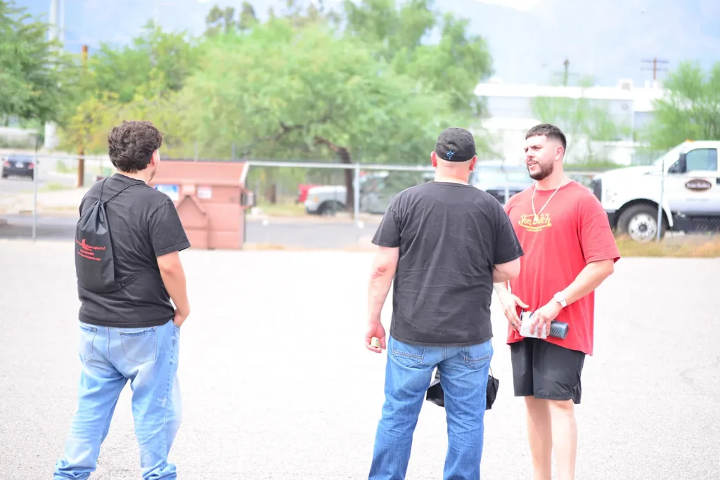 Three men chat in a sunny parking lot during a driver appreciation event, one carrying a Stevens Transport drawstring bag, with trees and a white work truck in the background.