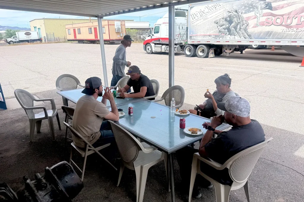 Students and staff eat burgers and drink sodas at patio tables under a covered area, with a Southwest Truck Driver Training semi truck and patriotic trailer parked in the yard behind them.