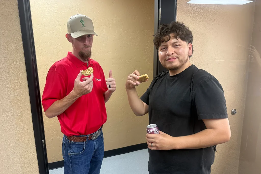 A Southwest Truck Driver Training instructor in a red polo and cactus logo cap gives a thumbs-up while eating a cookie beside a student holding a cookie and a can of soda.