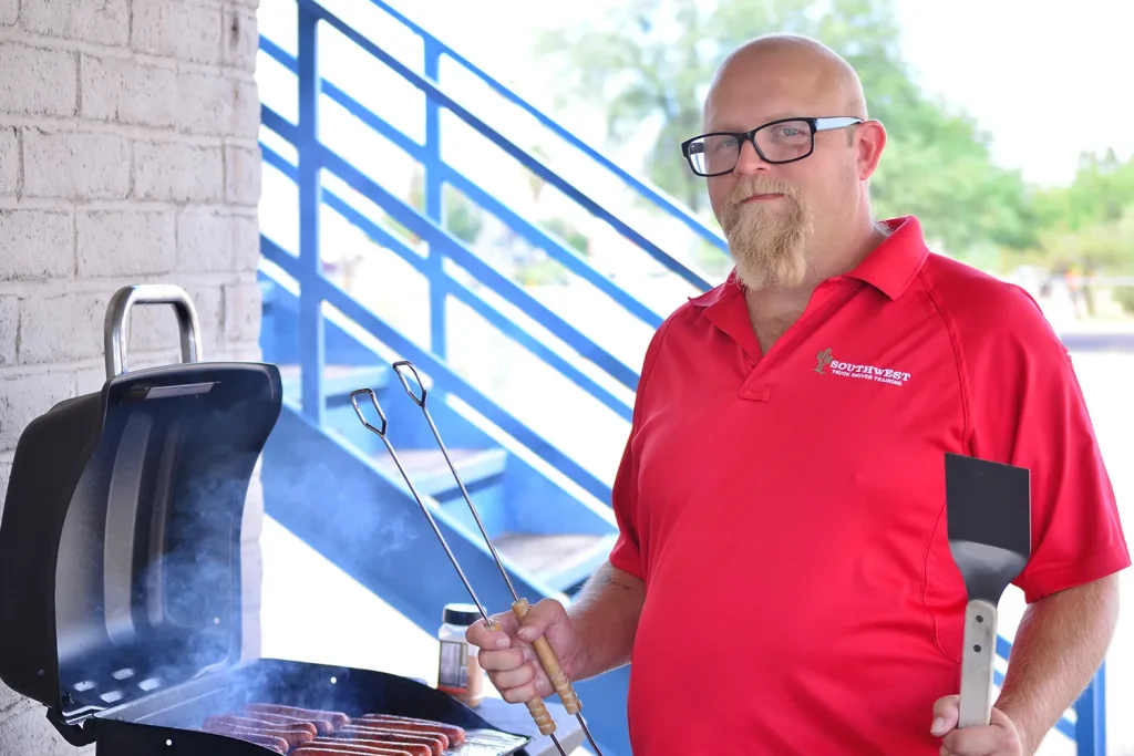 A Southwest Truck Driver Training staff member in a red polo and glasses holds grilling tongs and a spatula beside a grill of sizzling hot dogs next to blue exterior stairs.