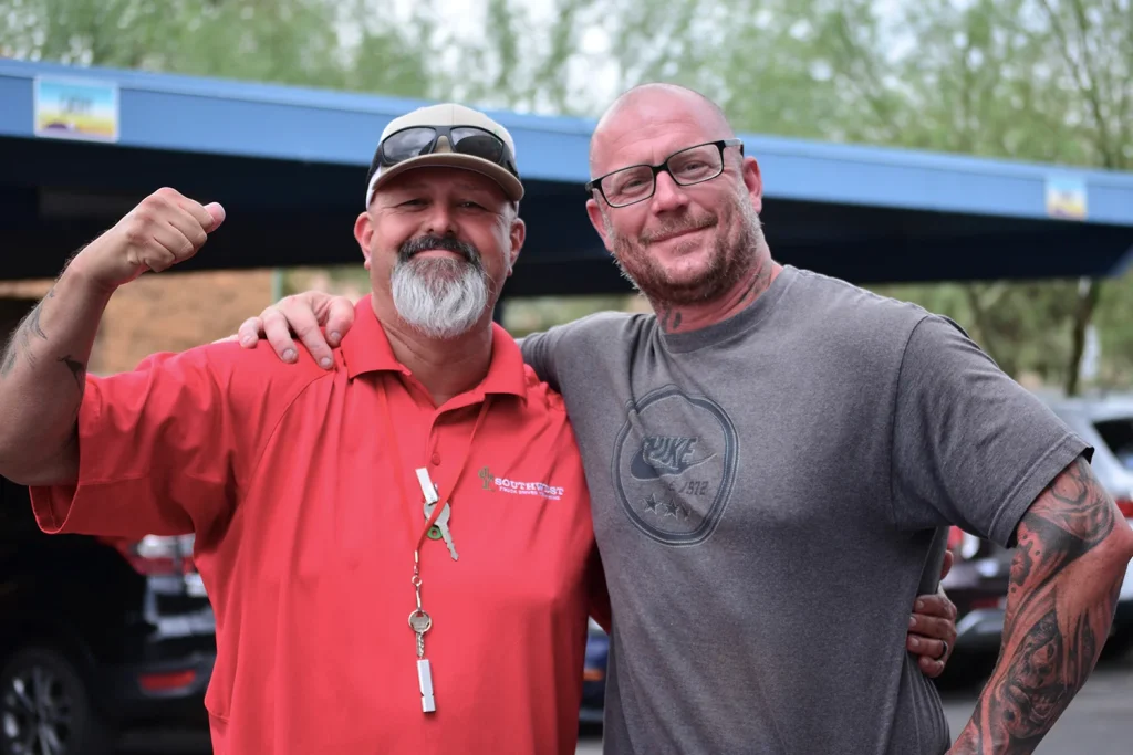 A smiling Southwest Truck Driver Training instructor in a red polo and cap flexes his arm while posing with his arm around a tattooed man in a gray T-shirt