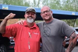 A smiling Southwest Truck Driver Training instructor in a red polo and cap flexes his arm while posing with his arm around a tattooed man in a gray T-shirt