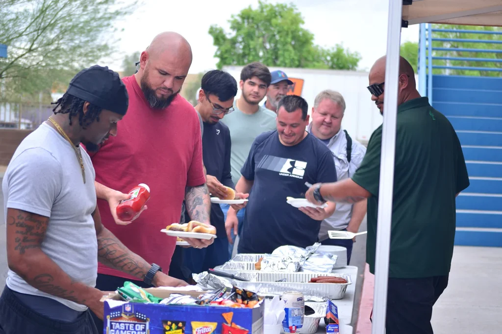 A line of students holding plates moves along a buffet table of foil trays, hot dogs, and a box of assorted chips while a staff member in a green polo serves food under a canopy.