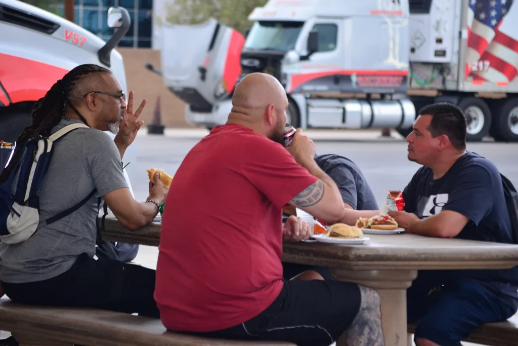Students sit at a stone picnic table eating hot dogs, burgers, and chips, with Southwest Truck Driver Training semi trucks parked in the background.