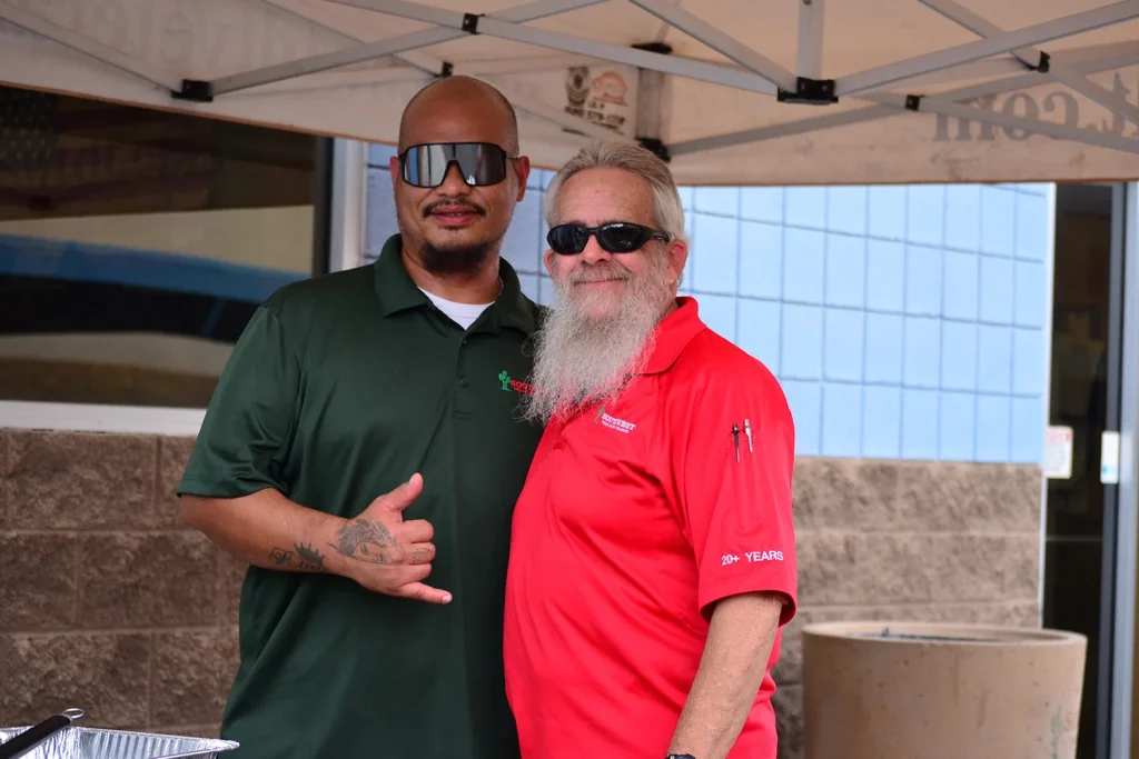 Two Southwest Truck Driver Training staff members pose under a canopy tent, one in a green polo flashing a shaka sign and the other, with a long white beard, in a red polo embroidered with "20+ Years."