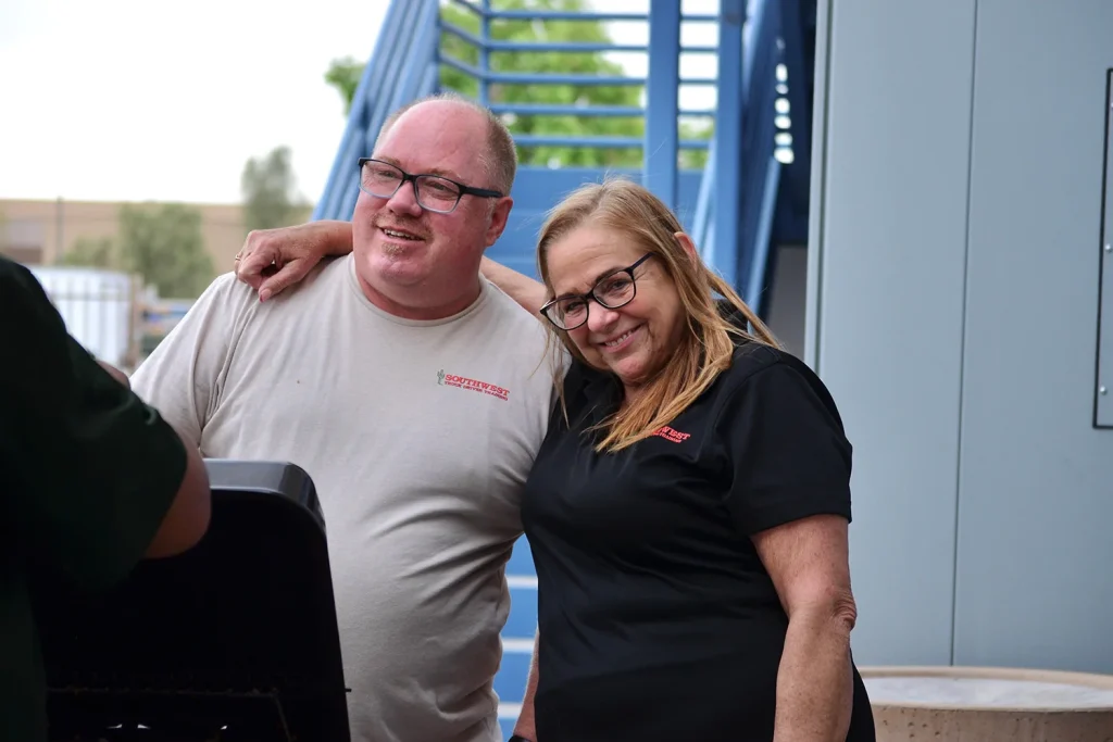 Two smiling Southwest Truck Driver Training staff members, a man in a tan T-shirt and a woman in a black polo, stand arm in arm beside a grill in front of blue exterior stairs.