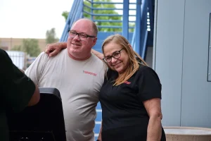 Two smiling Southwest Truck Driver Training staff members, a man in a tan T-shirt and a woman in a black polo, stand arm in arm beside a grill in front of blue exterior stairs.