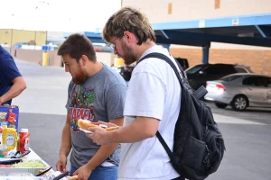 Two young men in casual T-shirts, one wearing a backpack and holding a plate with a hot dog, browse a buffet table stocked with chips, condiments, and shredded lettuce