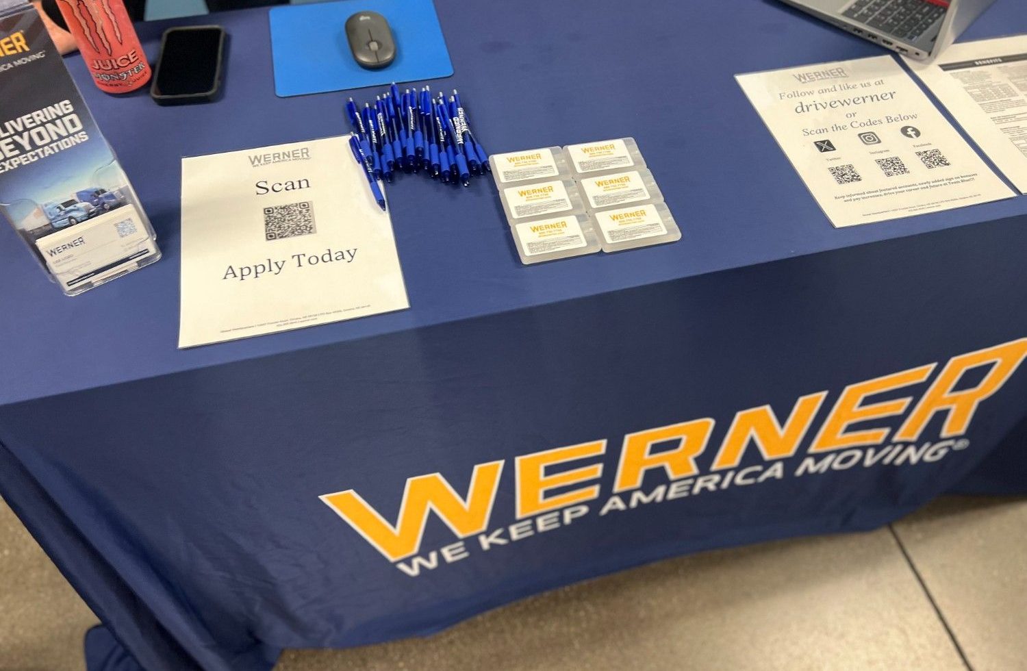 A Werner Enterprises recruiting table with a branded blue tablecloth reading "We Keep America Moving," set with an "Apply Today" QR code sign, pens, brochures, and promotional cards at a job fair.