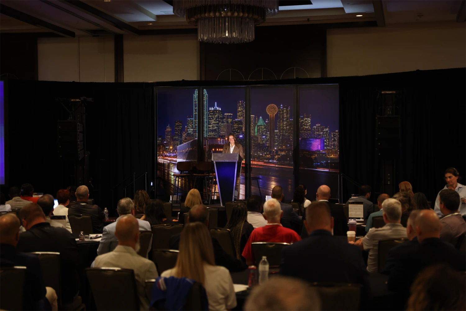 Samantha speaks at a podium on stage in front of a backdrop of the Dallas skyline at night, as a large audience listens at tables in a hotel ballroom.