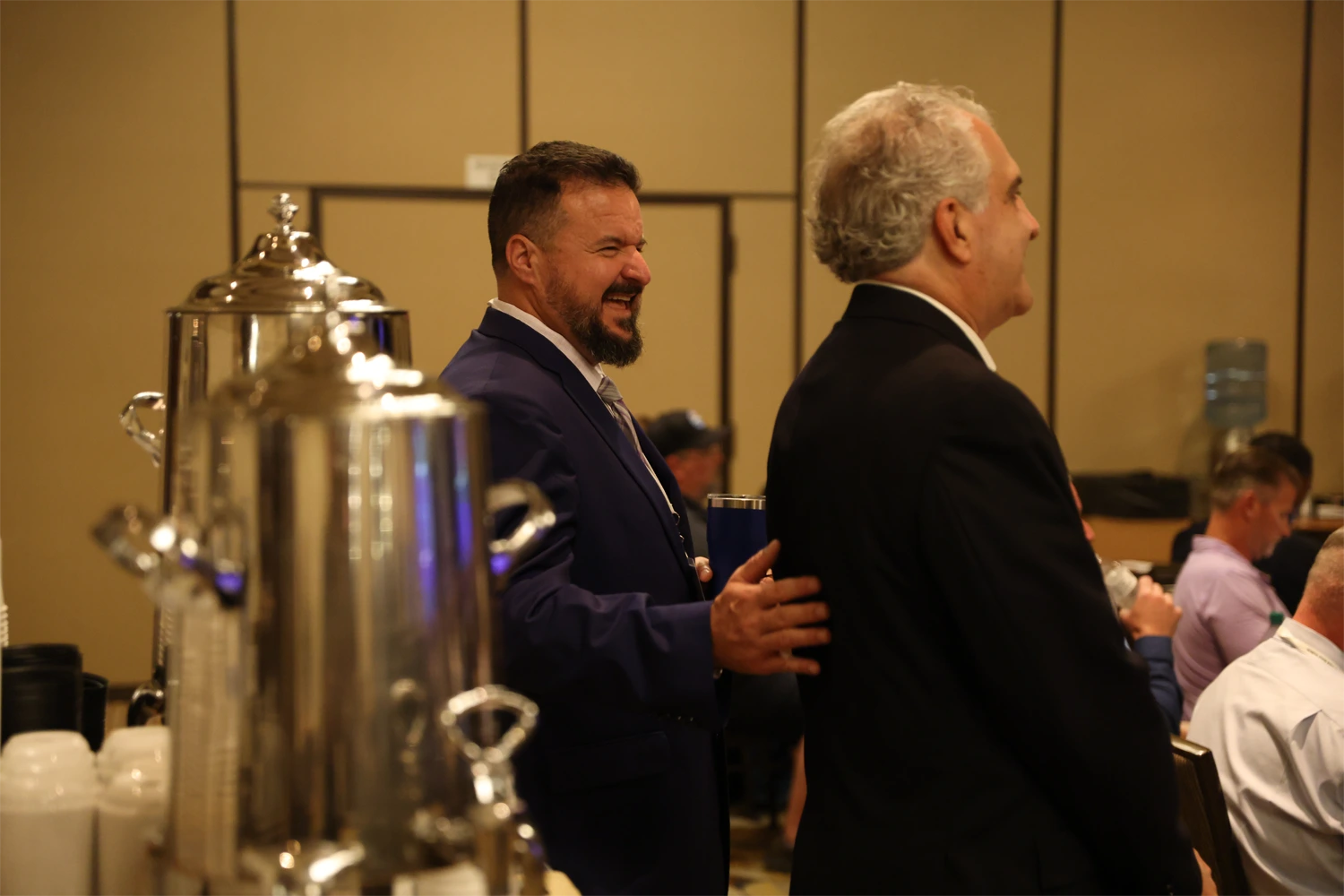 Sean in a navy suit pats Bruce in a dark suit on the back beside a coffee station at a conference event, with attendees seated at tables in the background.