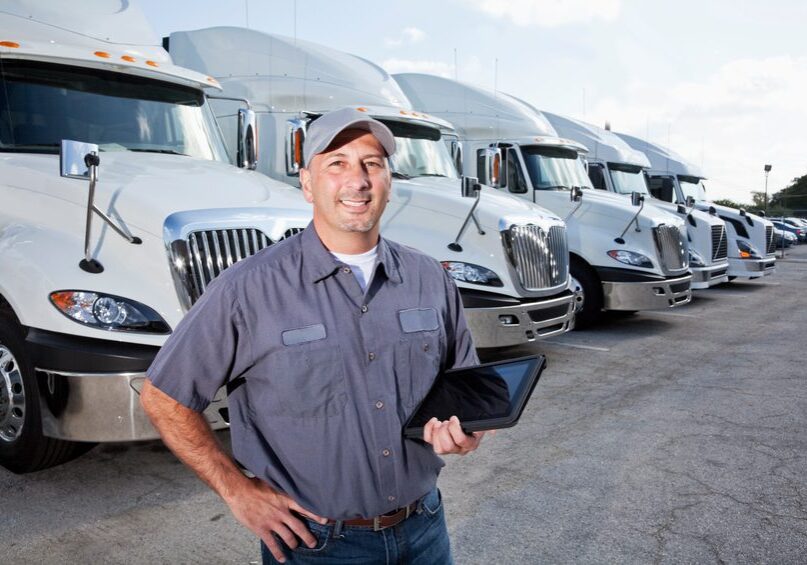A truck driver is standing in a large parking lot in front of several large trucks.  He is facing the camera, and he is wearing a short-sleeved blue shirt, blue jeans, a brown belt and a gray hat.  He is holding a black tablet with his left hand.  There are six big rigs seen lined up behind him, going from left to right.  The large trucks all look the same, being all white and having silver grills and mirrors.  There are multiple cars seen parked to the right of the trucks.  A cloudy blue sky is seen in the background.